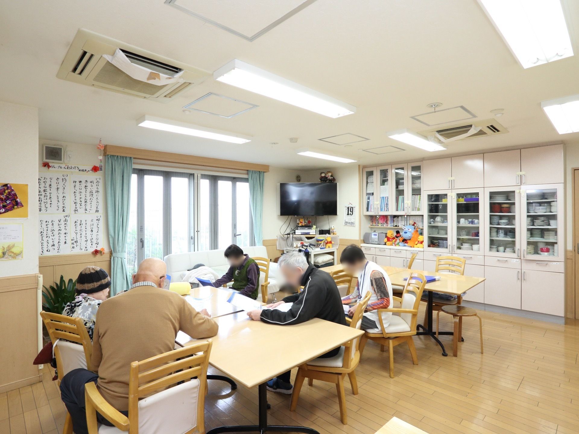 椅子に座り、入居者様やスタッフが紙を読んでいる。大型テレビや、食器棚が配置されている。観葉植物が飾られている。