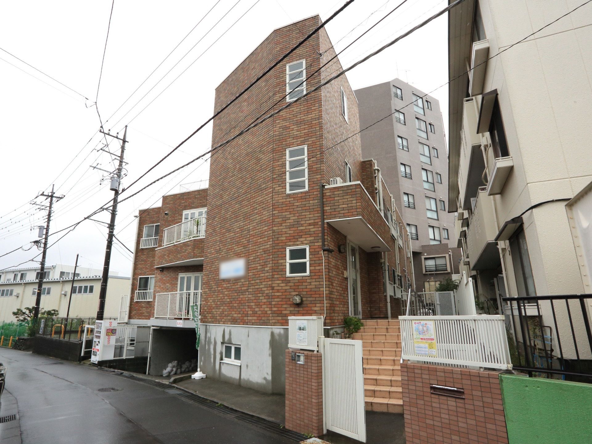 レンガ風の外壁の建物で、隣に飲料の自動販売機がある。建物下に駐車場がある。入り口前に手すり付きの階段が設置してある。