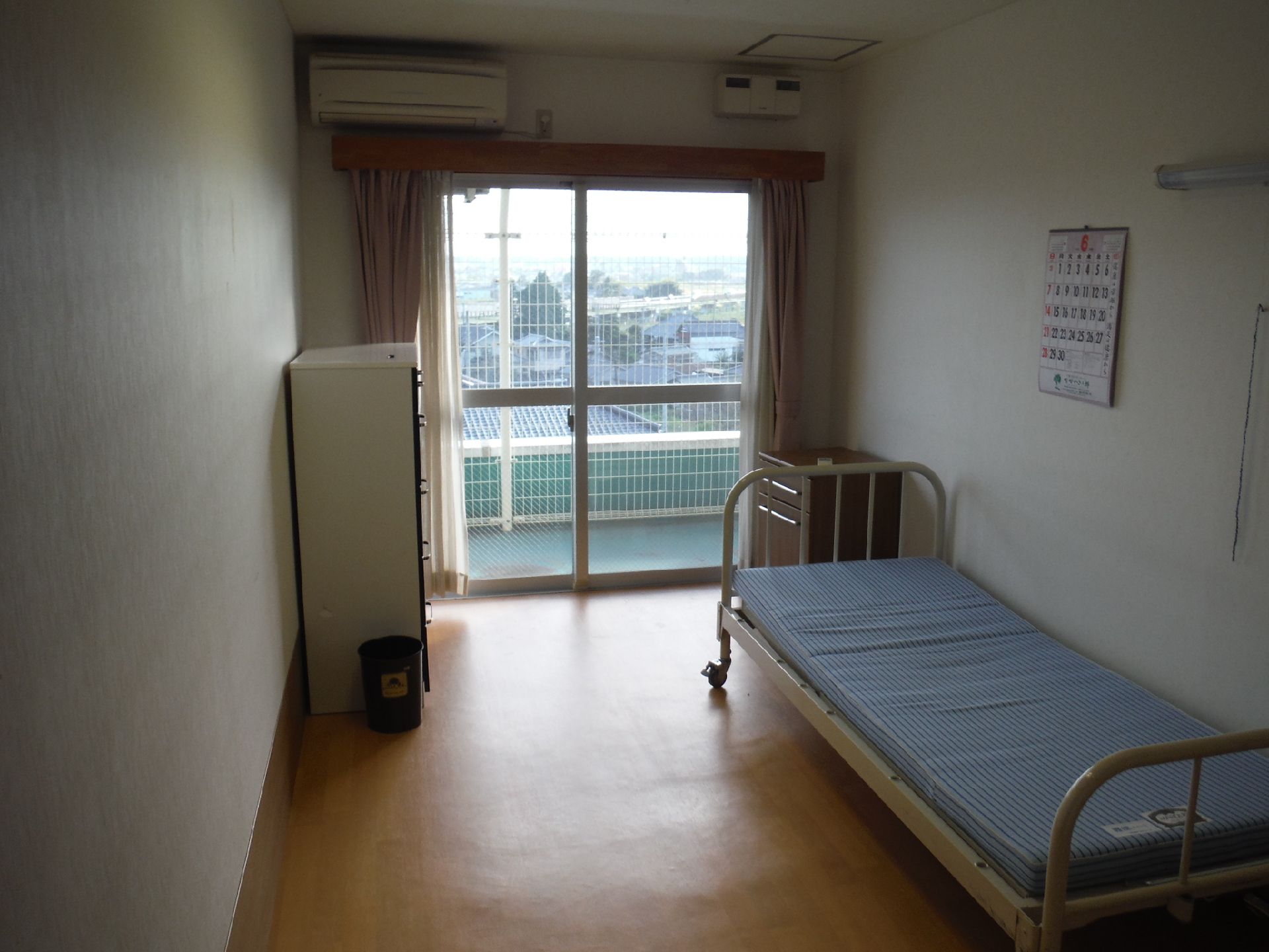 小さな冷蔵庫が置かれた部屋。反対側にはパイプベッドがあり、青と白の縞模様のうすいマットレスが乗っている。