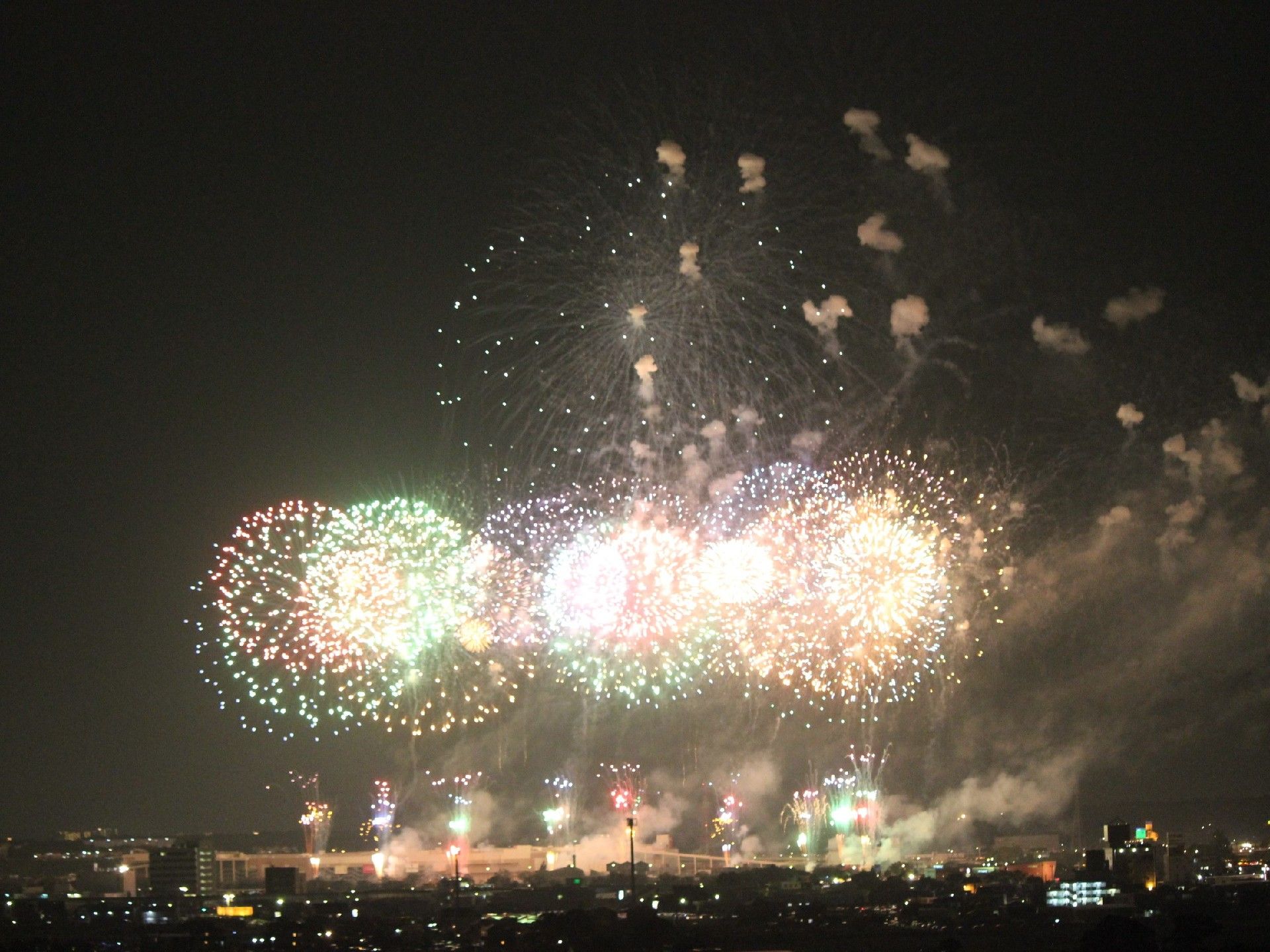 夜空にいくつもの花火が打ち上げられている。色とりどりできれいな円形になっているものや上から落ちてくるものなど形もさまざま。