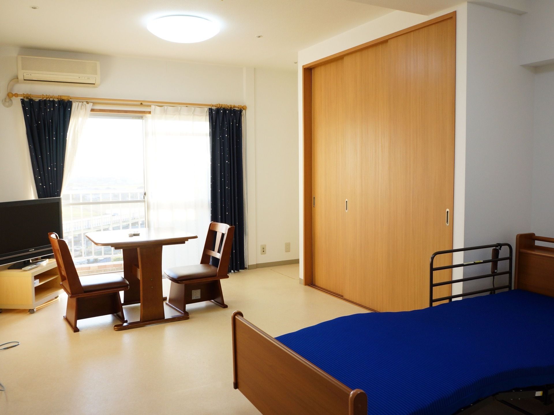 介護用の電動ベッドが完備されている。窓があり、そばにはテレビが設置されている。その前にはテーブルと椅子が２脚置いてある。