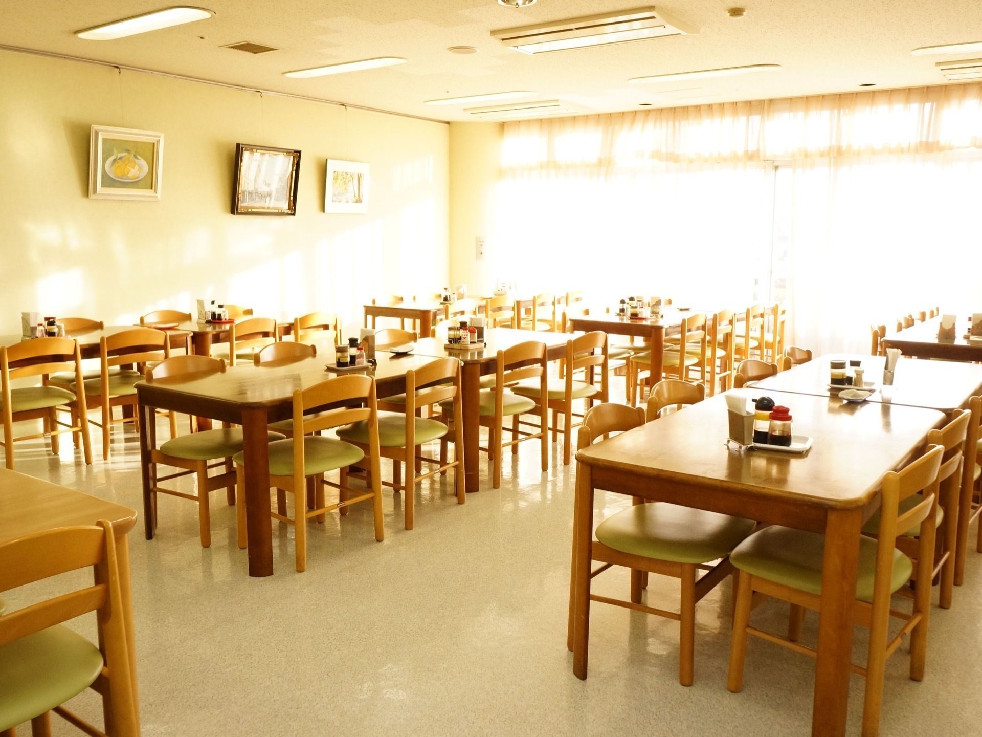 部屋には大きな窓がある。たくさんのテーブルと椅子が並べられている。テーブルの上には調味料とナプキンが置かれている。