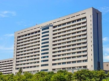 ベージュ色の高層階のマンションのような建物が並んでいる。建物の前には、緑の葉が生い茂る木々が連なっている。