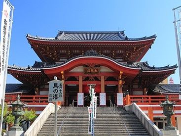 黒色の瓦屋根、朱色の柱からなる寺社である。寺社ののぼりが複数立っている。馬の塔と記された看板がある。