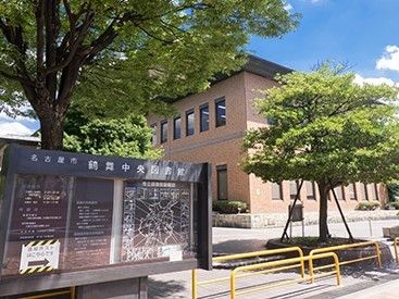 名古屋市の図書館である。淡い茶色のレンガ造りの外観で、周囲には緑の葉が生い茂る植栽がある。茶色の大きな掲示板には掲示物がある。