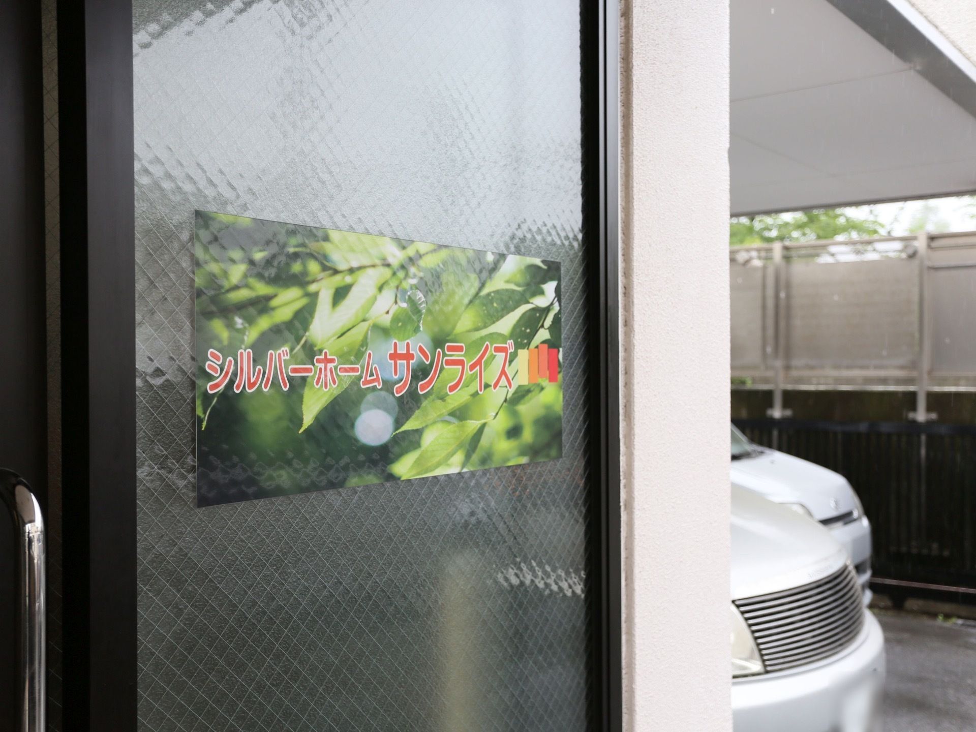 玄関扉はすりガラスでできており、目の高さには緑の枝葉をバックに、施設名のロゴが明るいオレンジ色で印刷された紙が貼られている。
