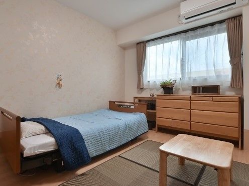 居室には窓がありエアコンが設置されている。床はフローリングで、ベッド、タンス、敷物とテーブルが置かれている。