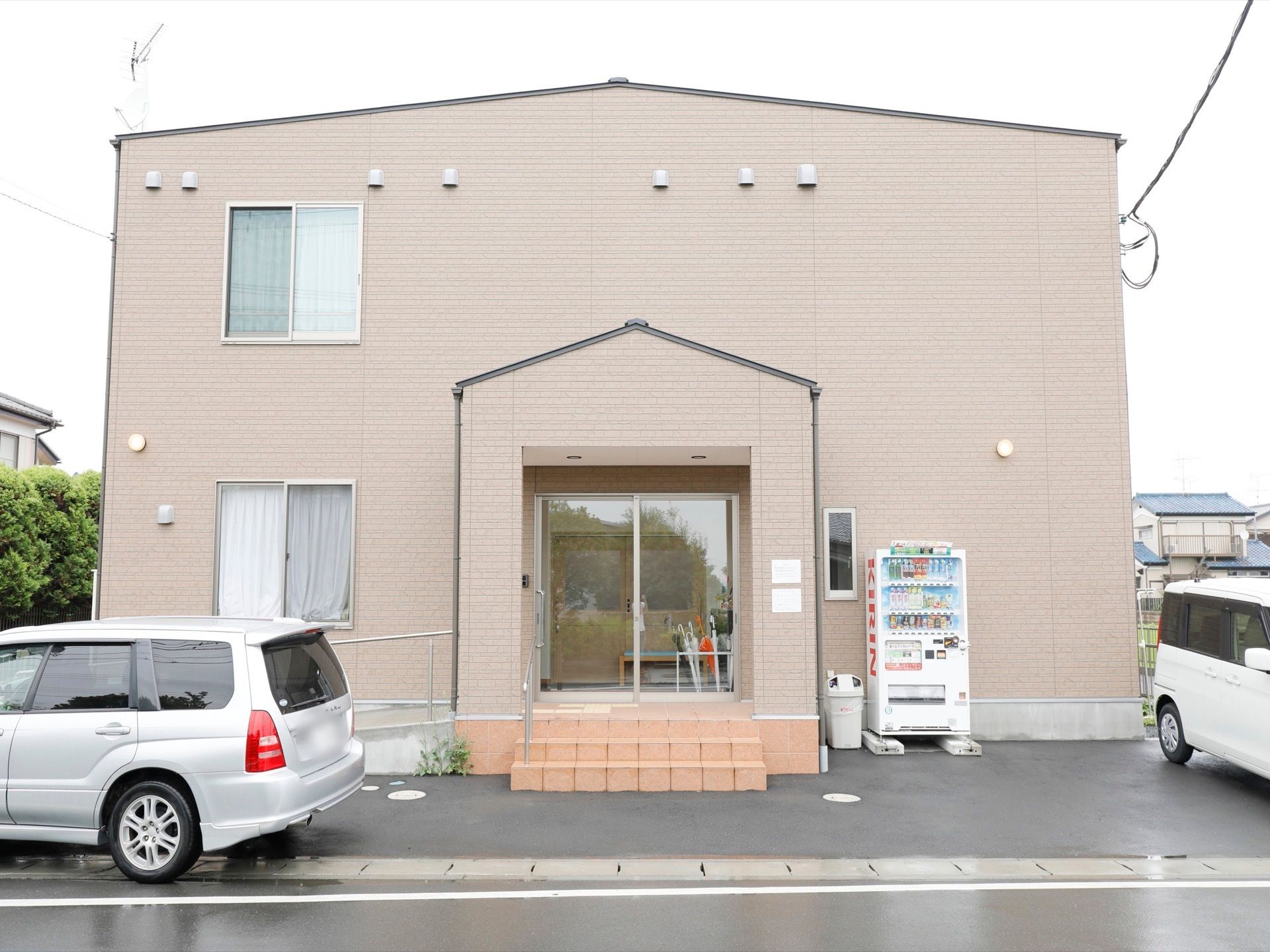 木造２階建てのグレーの建物の玄関正面に自動販売機がある。入口へのアプローチは、階段とスロープがあり手すりがついている。