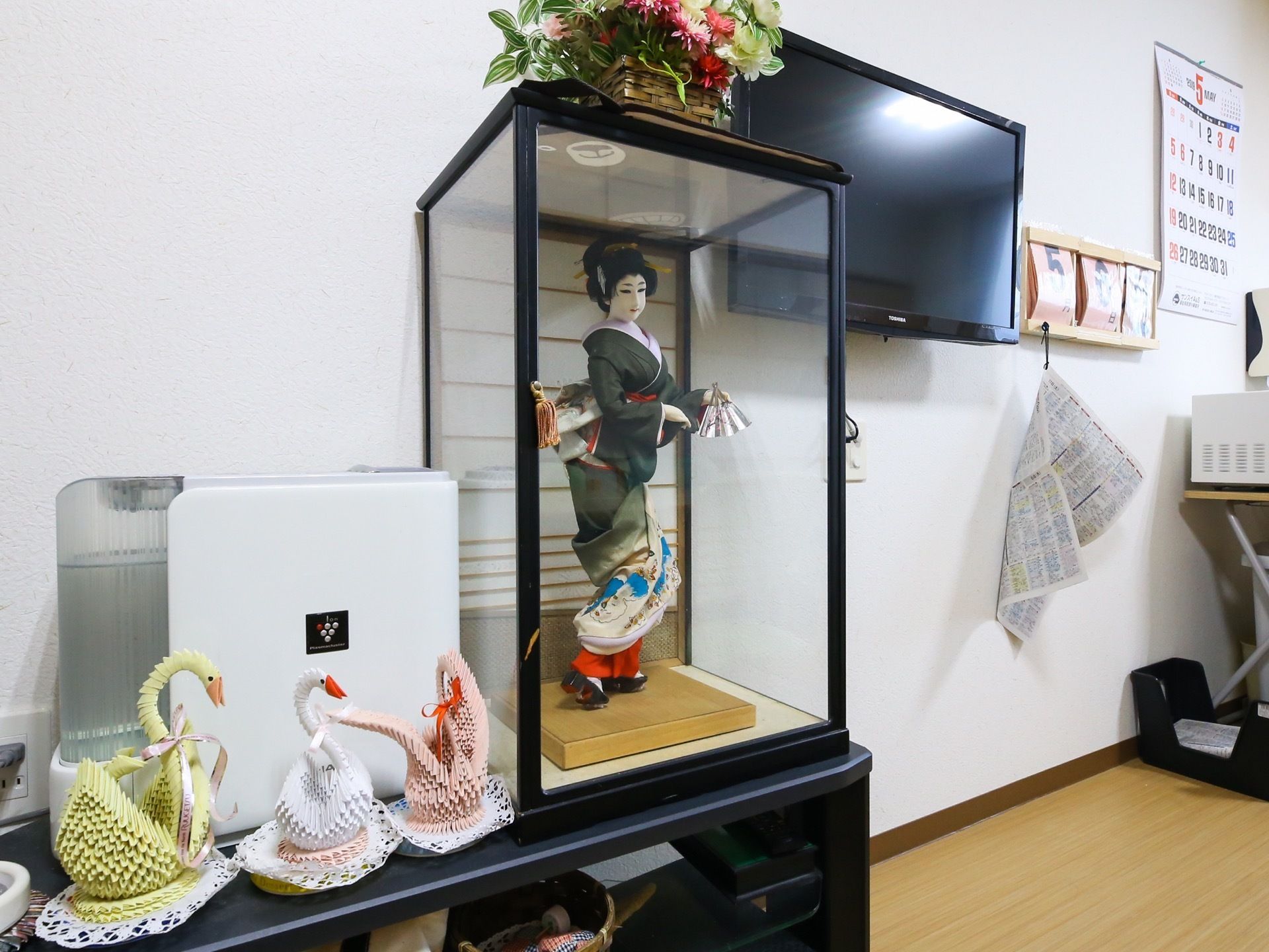 壁にテレビやカレンダーが掛けられている。その下にケースに入った和装の人形や加湿器が置かれた棚がある。