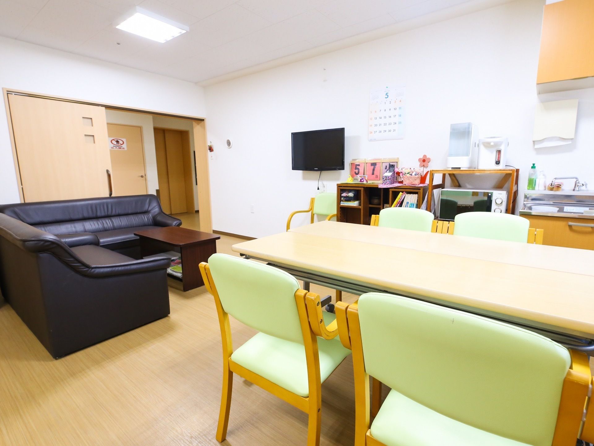 フローリングの部屋に、ダイニングテーブルとソファが置かれている。キッチンがあり、電子レンジやポットが設置されている。