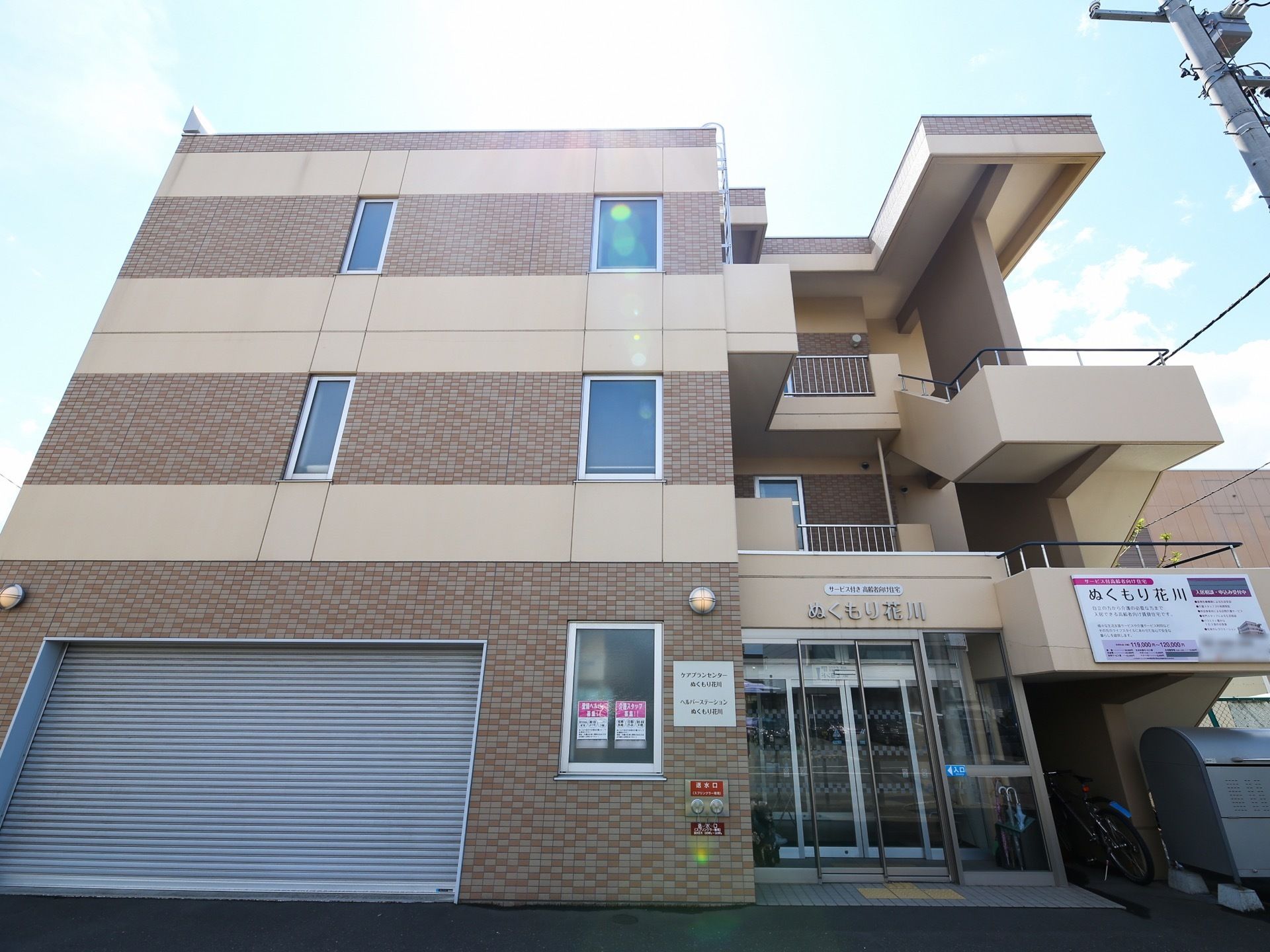 マンションのようなタイル張りの柔らかい色味の建物がある。玄関は自動ドアになっていて、その横はシャッター付きのガレージになっている。