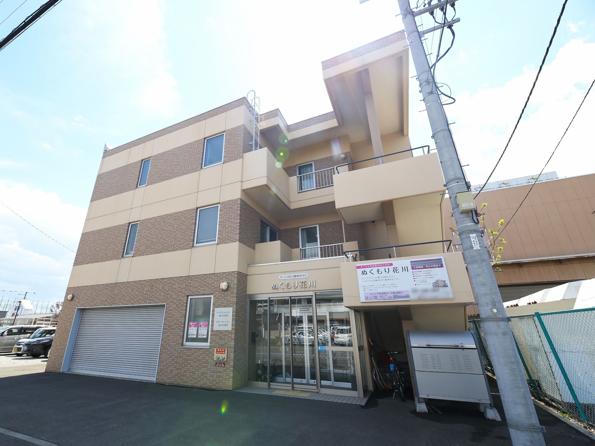 茶色のタイルとベージュの壁が印象的なマンションタイプの建物が建っている。１階部分はガレージになっていて、シャッターが下りている。