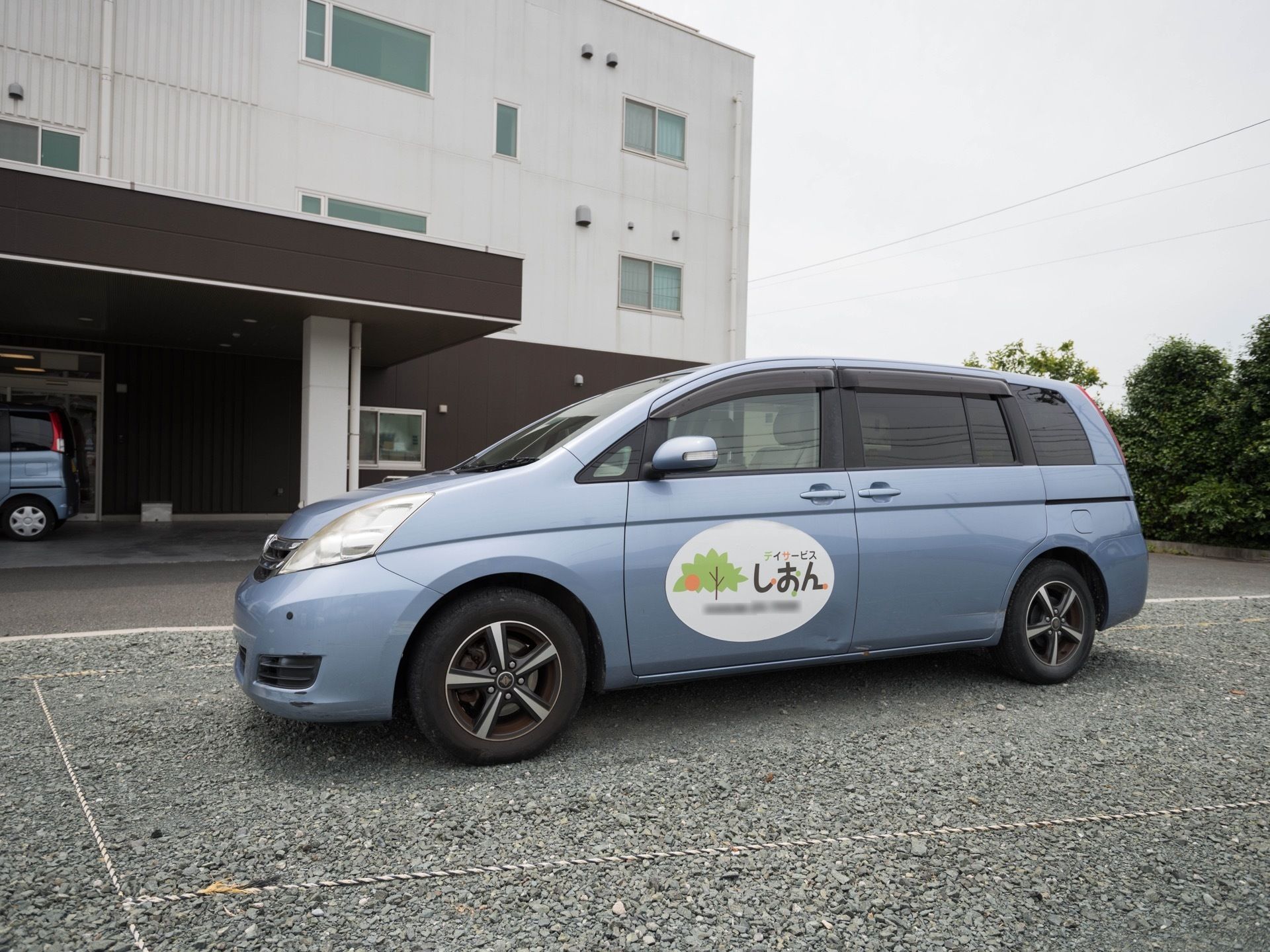 白色と茶色の３階建ての建物が建っている。敷地内に停められた水色の車の前扉にデイサービスの名称が表示されている。