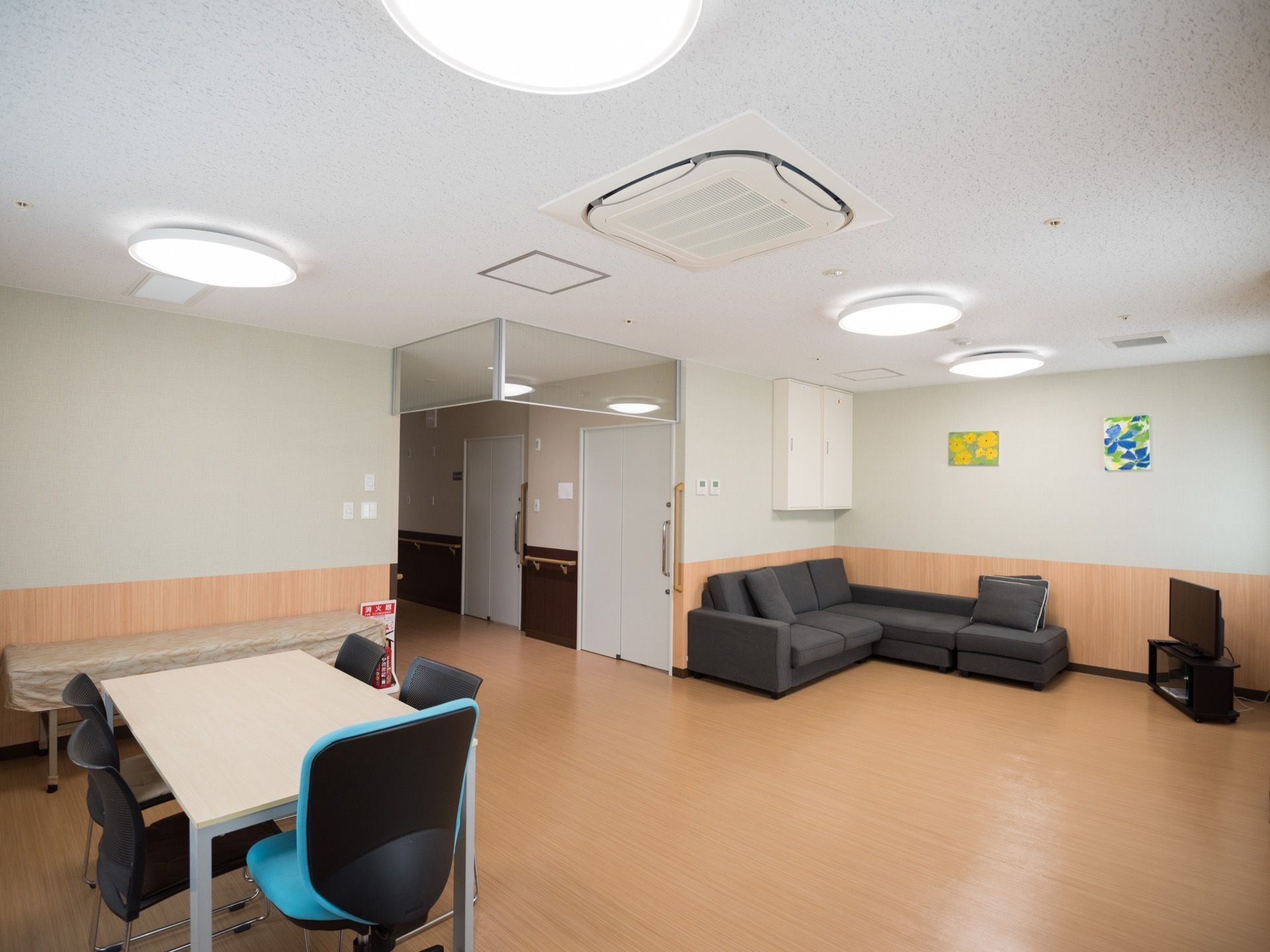 部屋の角に濃い灰色のソファーが置いてあり、その前に薄型テレビが設置されている。壁に２種類の絵が飾られている。