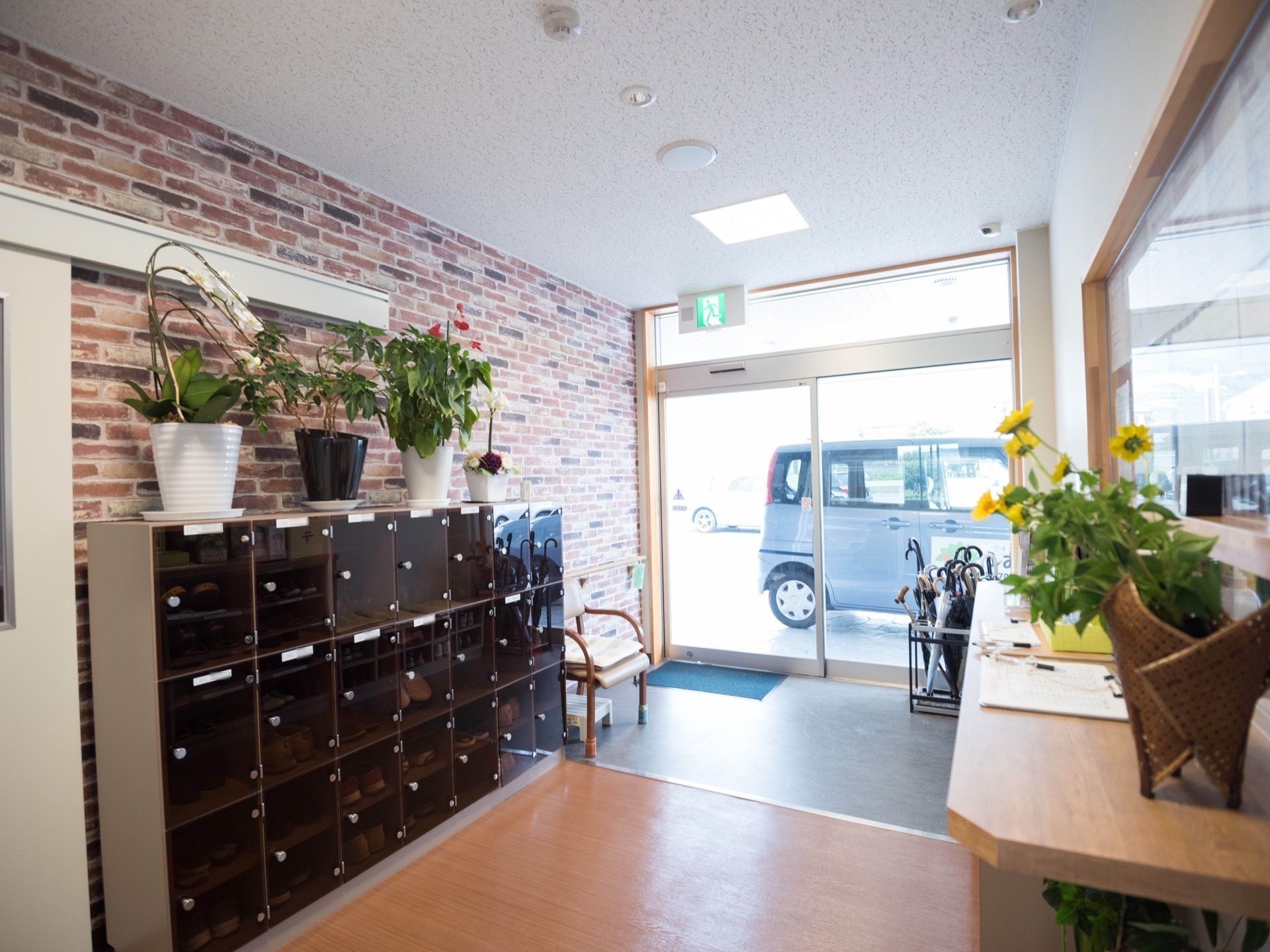 壁にレンガ調の壁紙が貼られている。茶色い透けた扉が付いた靴箱があり、その上にさまざまな花が置かれている。