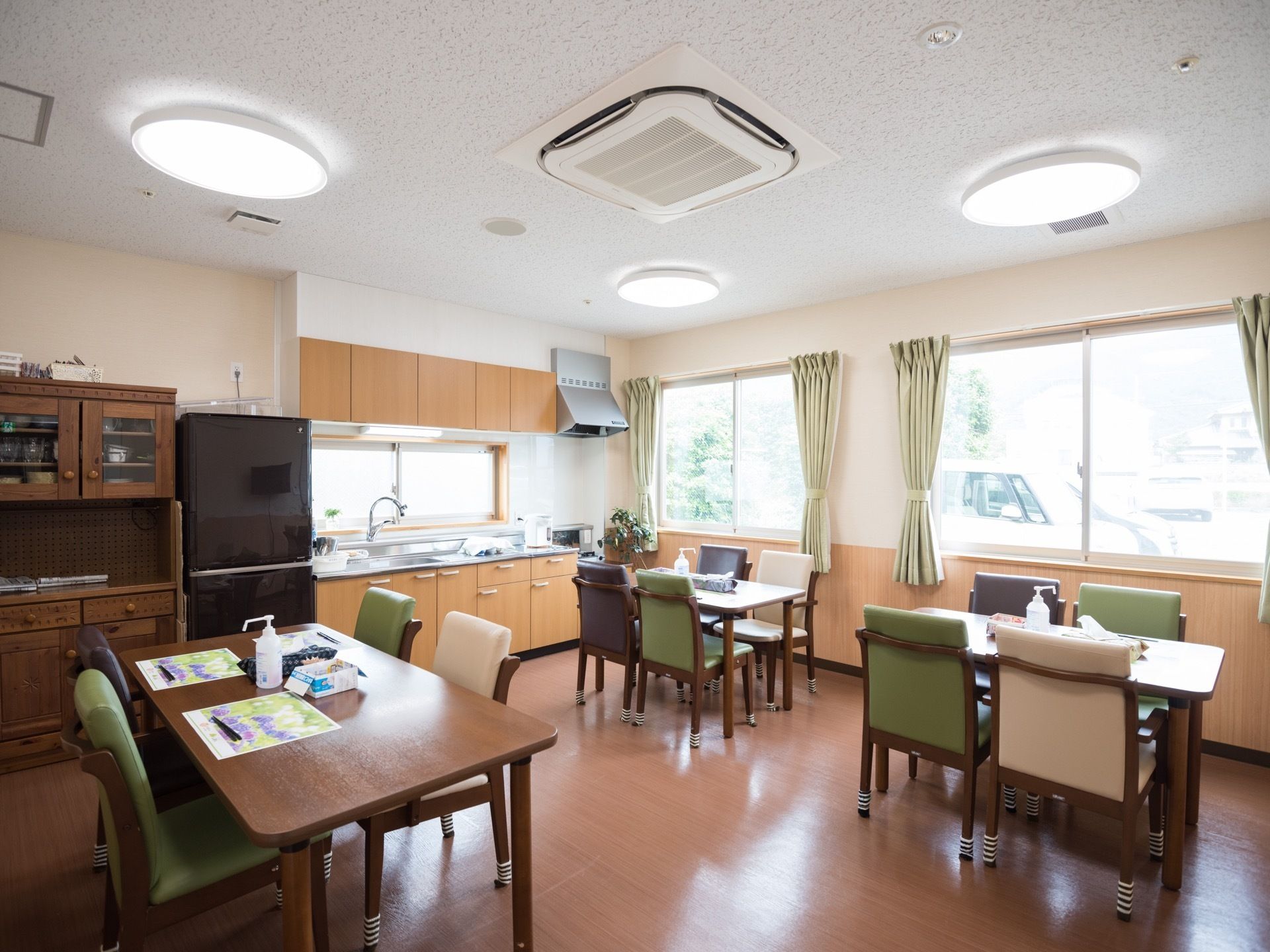 木目の扉のシステムキッチンがあり、その隣に冷蔵庫と食器棚が置いてある。天井のシーリングライトが点灯している。
