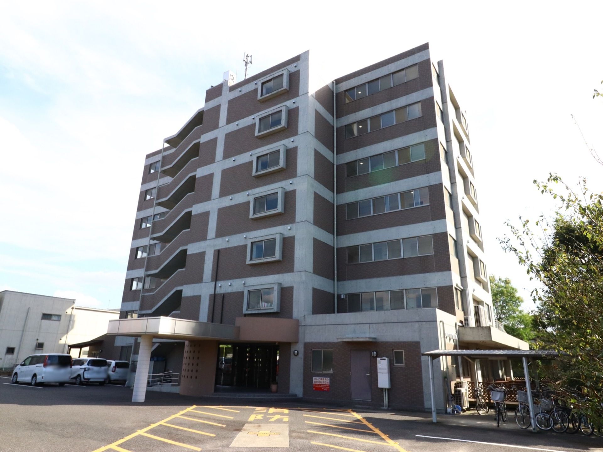 施設は７階建ての落ち着いた色の建物。建物の前には屋根付きの駐輪場や複数台停められる駐車場が用意されている。