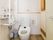 可動式の手すりが設置されたタンク付きの洋式トイレがある。壁に洗面台とペーパーホルダー、温水洗浄便座のリモコンが取り付けられている。