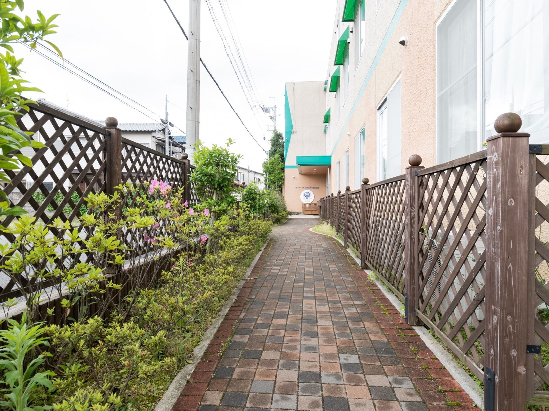 施設の脇から続く通路が、出入り口まで続いている。通路の両脇には、背の高い木製の柵が地面に固定される形で設置されている。