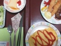 食事が配膳されている画像。ケチャップがかかったオムライス、エビフライ、ハンバーグ、ウインナーが盛り付けてあるお皿のほか、デザートが付いている。