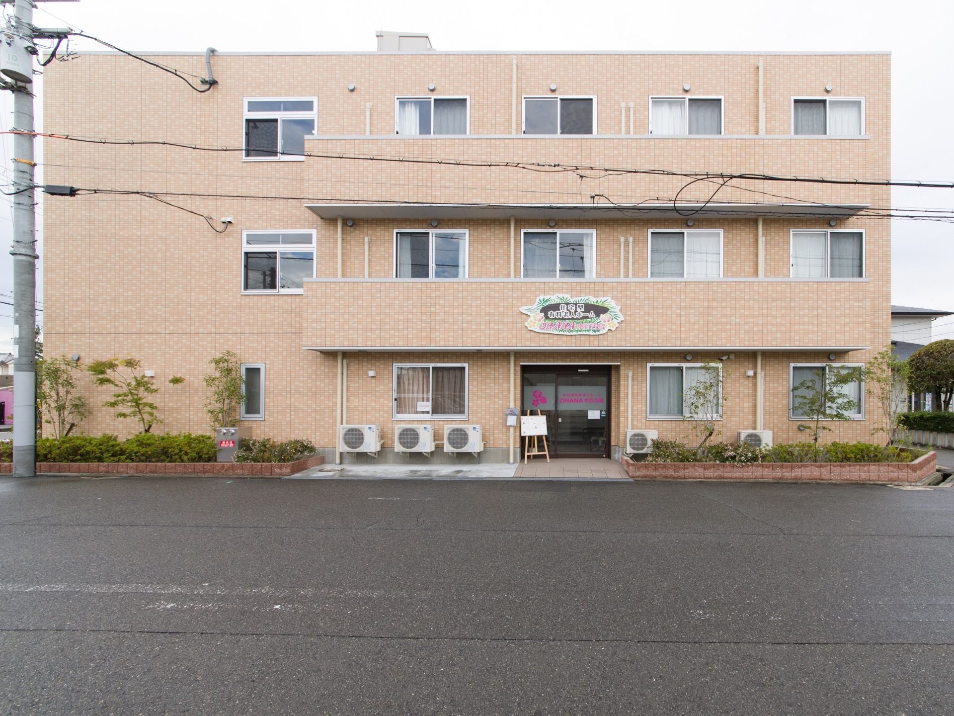 施設の正面外観である。3階建ての建物で、正面に室外機が複数台置かれている。周りの花壇には木や植物が植えてある。