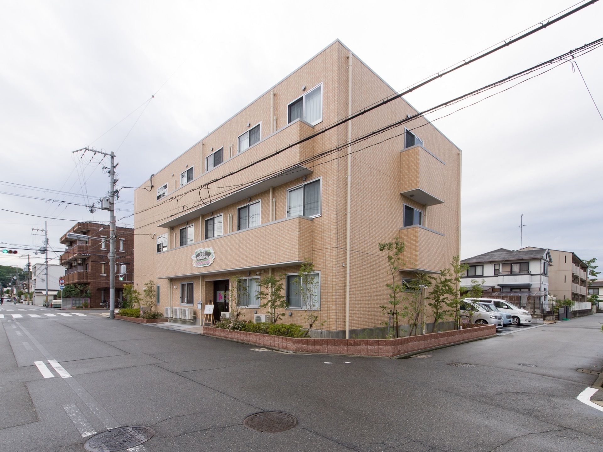 施設の外観の様子。3階建ての建物で建物の周囲には花壇が作られ立木が植えられている。施設裏には駐車場がある。