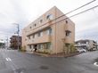 施設の外観の様子。3階建ての建物で建物の周囲には花壇が作られ立木が植えられている。施設裏には駐車場がある。