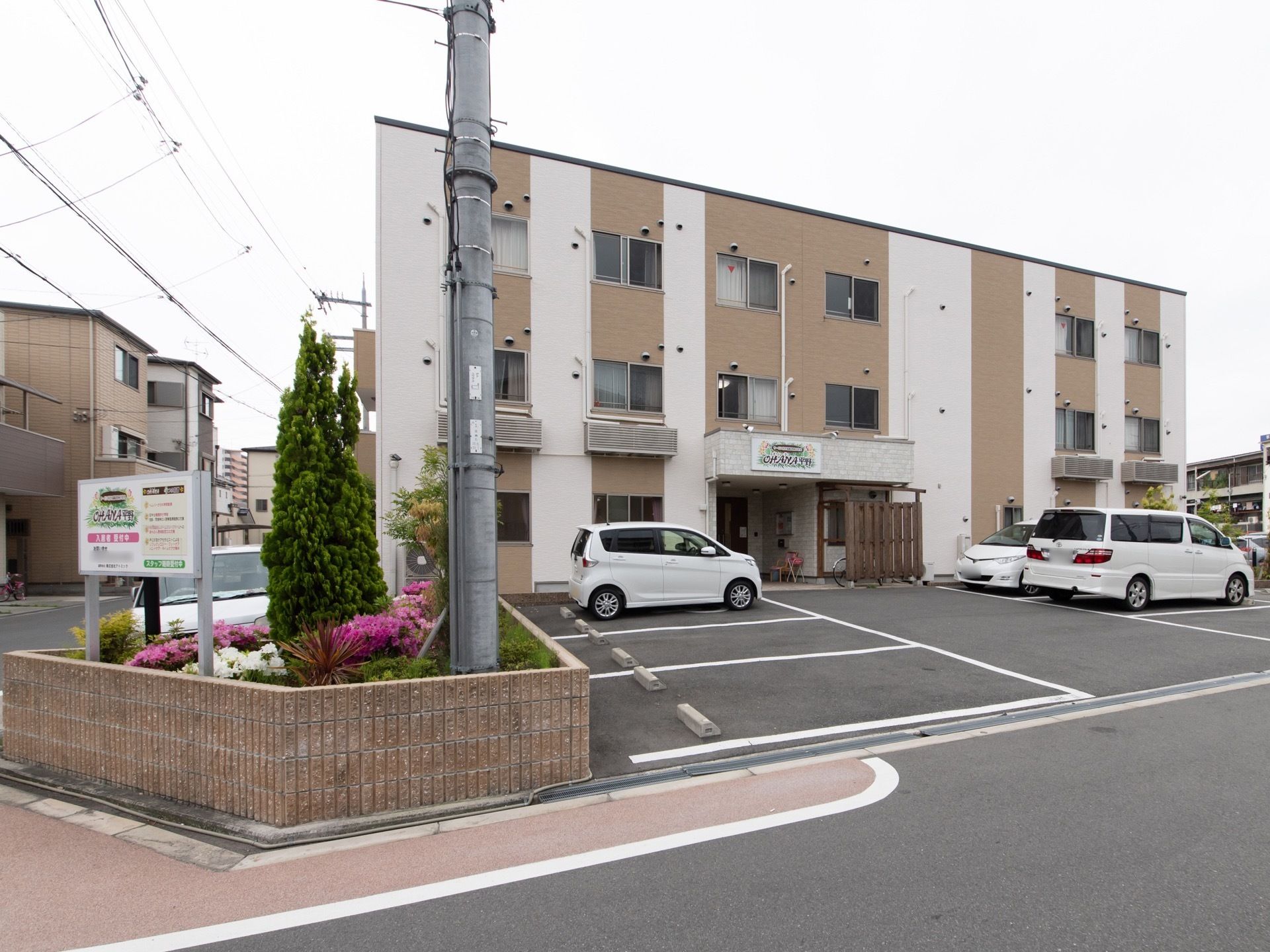 当施設は角地に立地しており、施設名を表示した看板を立てている。白と茶色のツートンの外壁の建物が目印になっている。