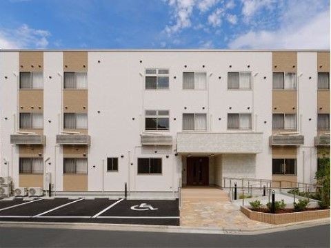 建物の前に駐車スペースがあり、玄関前にスロープと階段が設置された３階建ての施設