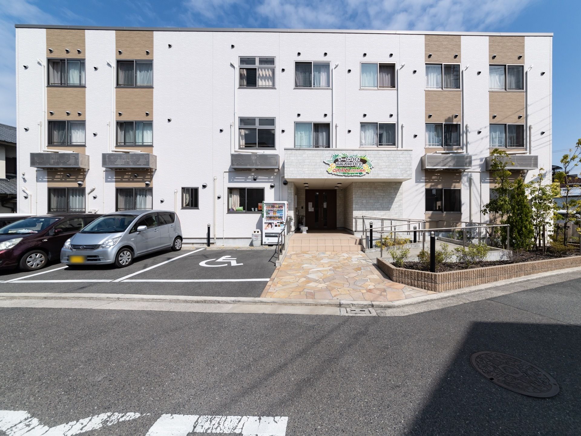 白色とベージュ色の２色が使われた落ち着いた雰囲気がある建物である。建物前の駐車場には車椅子をご利用の方専用のスペースを確保している。