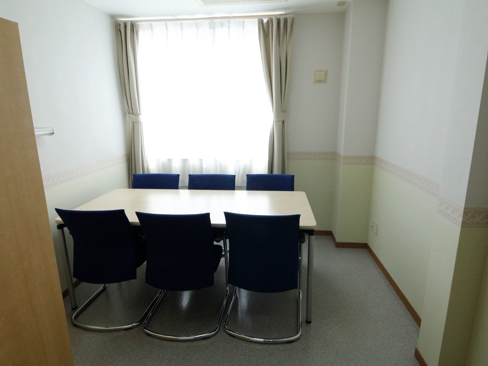 個室内に、６人掛けのテーブルが置かれている。座面の青い椅子が並んでいる。大きな窓が付いているので、明るい。