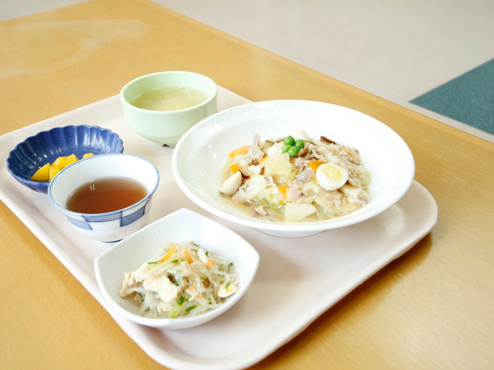 白色のトレイの上に具沢山の丼とスープに小鉢のおかずとフルーツ、そしてお茶を添えられた食事である。食材は、食べやすい大きさにカットしている。