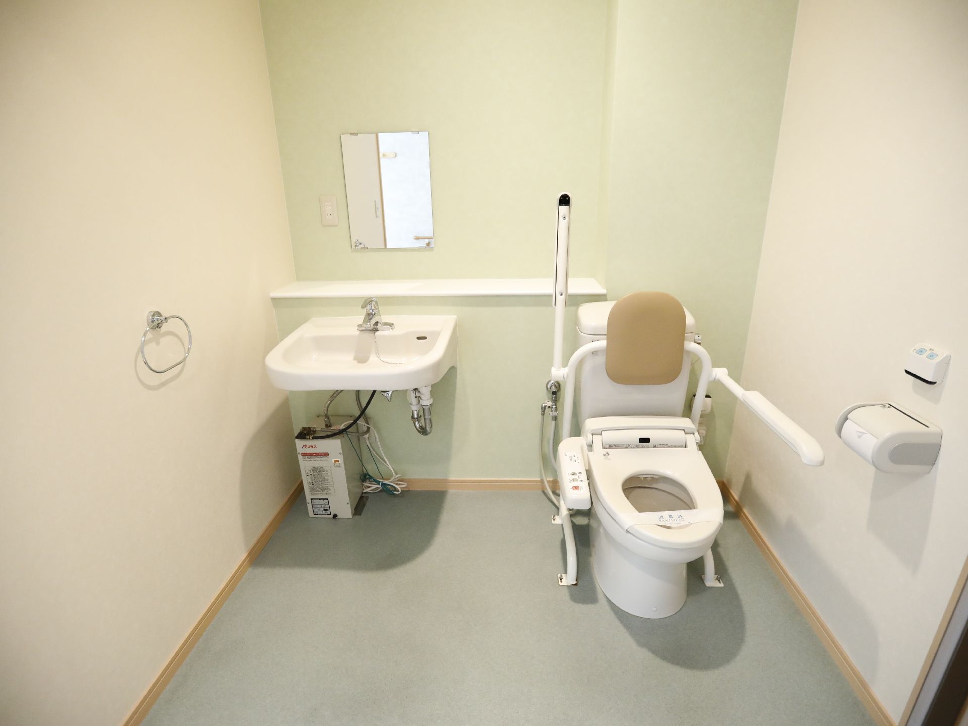 背あてと可動式のひじ掛けが付いた洋式トイレが設置されている。隣には洗面台が置かれ、正面には鏡が付いている。
