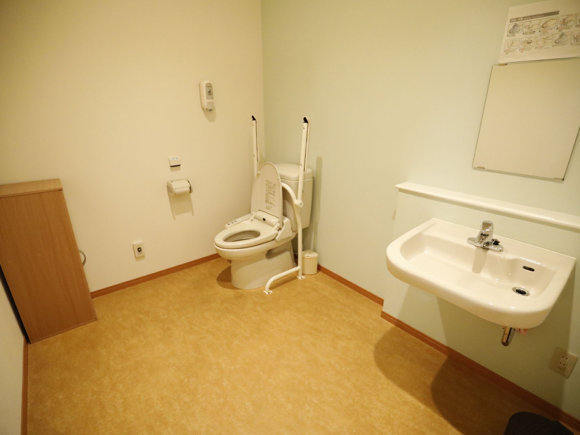 洋式トイレの向かいに、収納棚がある。トイレ清掃用のブラシがある。手洗い場がある。手の洗い方の張り紙がしてある。