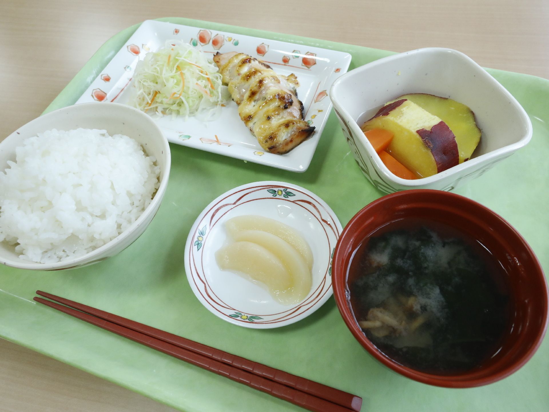 小さな小鉢に野菜が盛り付けてある。すべてのおかずが緑色のトレイにのっている。千切りされたキャベツが角皿に盛ってある。フルーツもついている。