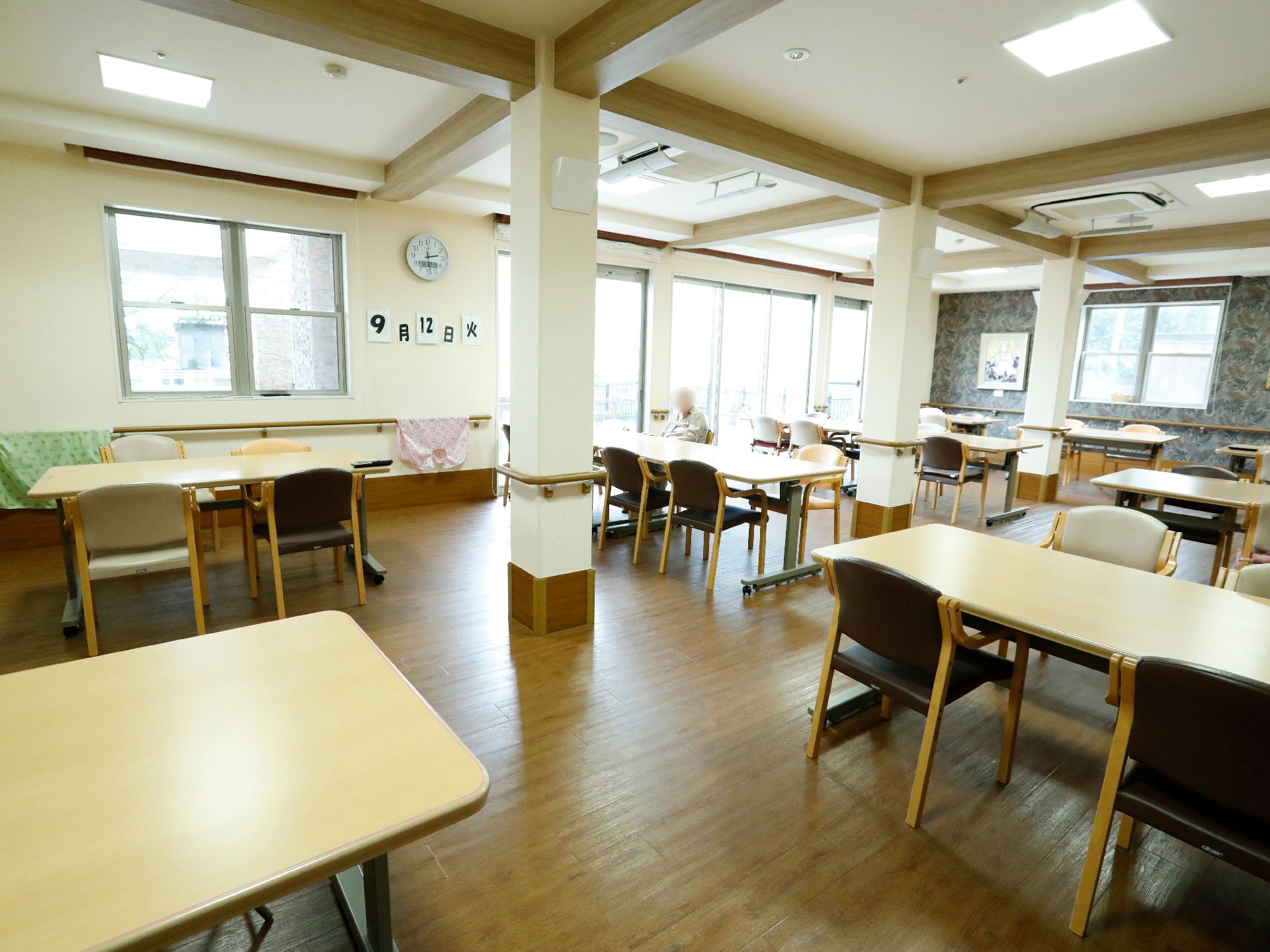 食堂には複数のテーブルと椅子がゆとりをもって配置されている。大きな窓があり、壁には手すりが付いている。