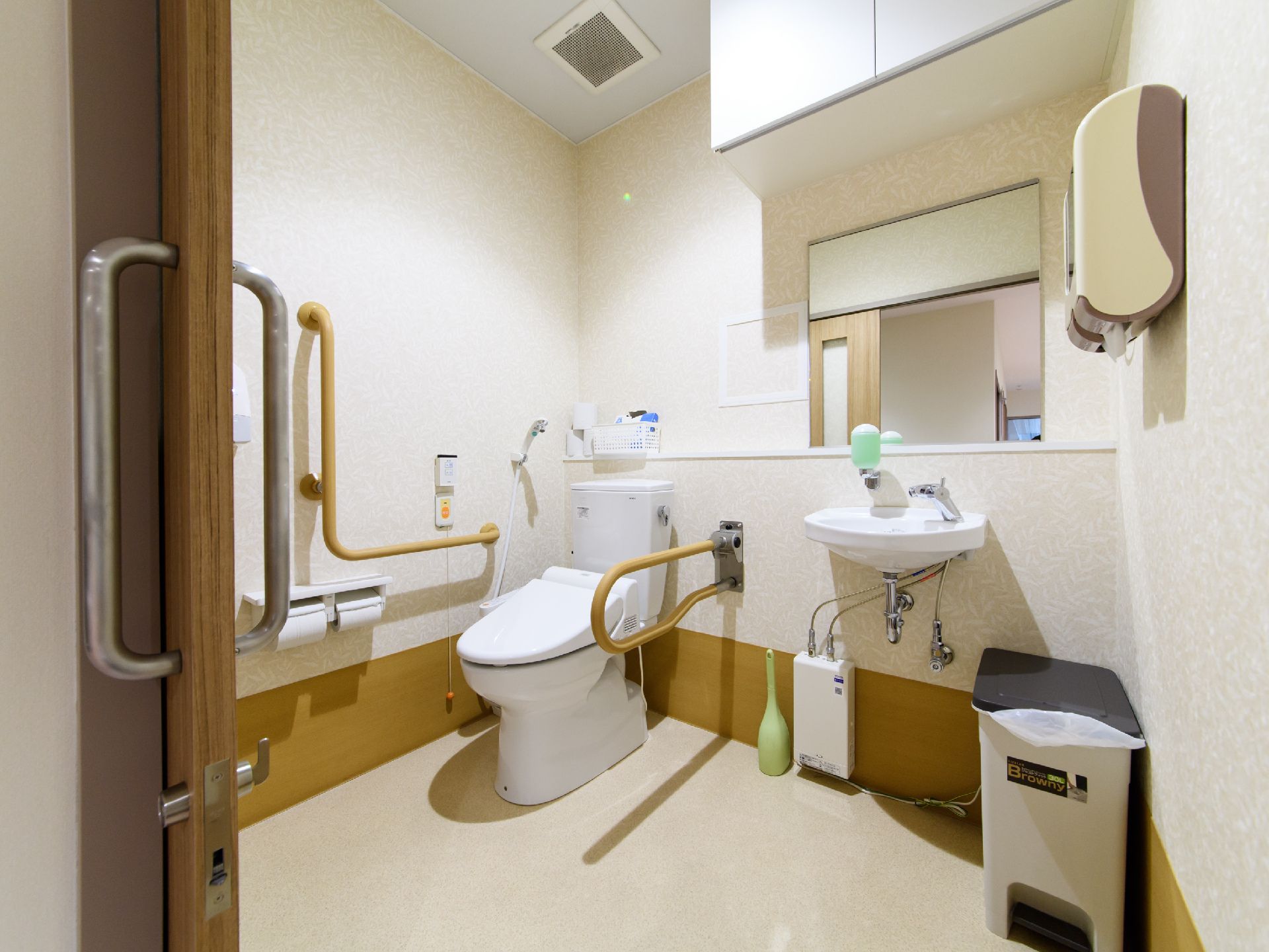 白色基調のデザイン仕様の壁に薄い茶色の床のトイレである。洗面台が設置してあり、壁には茶色の手すりが取り付けてある。