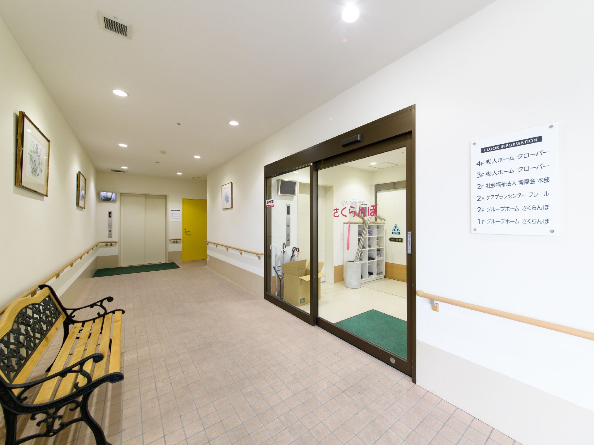 白色の天井と壁に薄いピンク色のタイル調の床である。奥の正面に白色のドアのエレベーターがある。壁際にお洒落なベンチが置いてある。