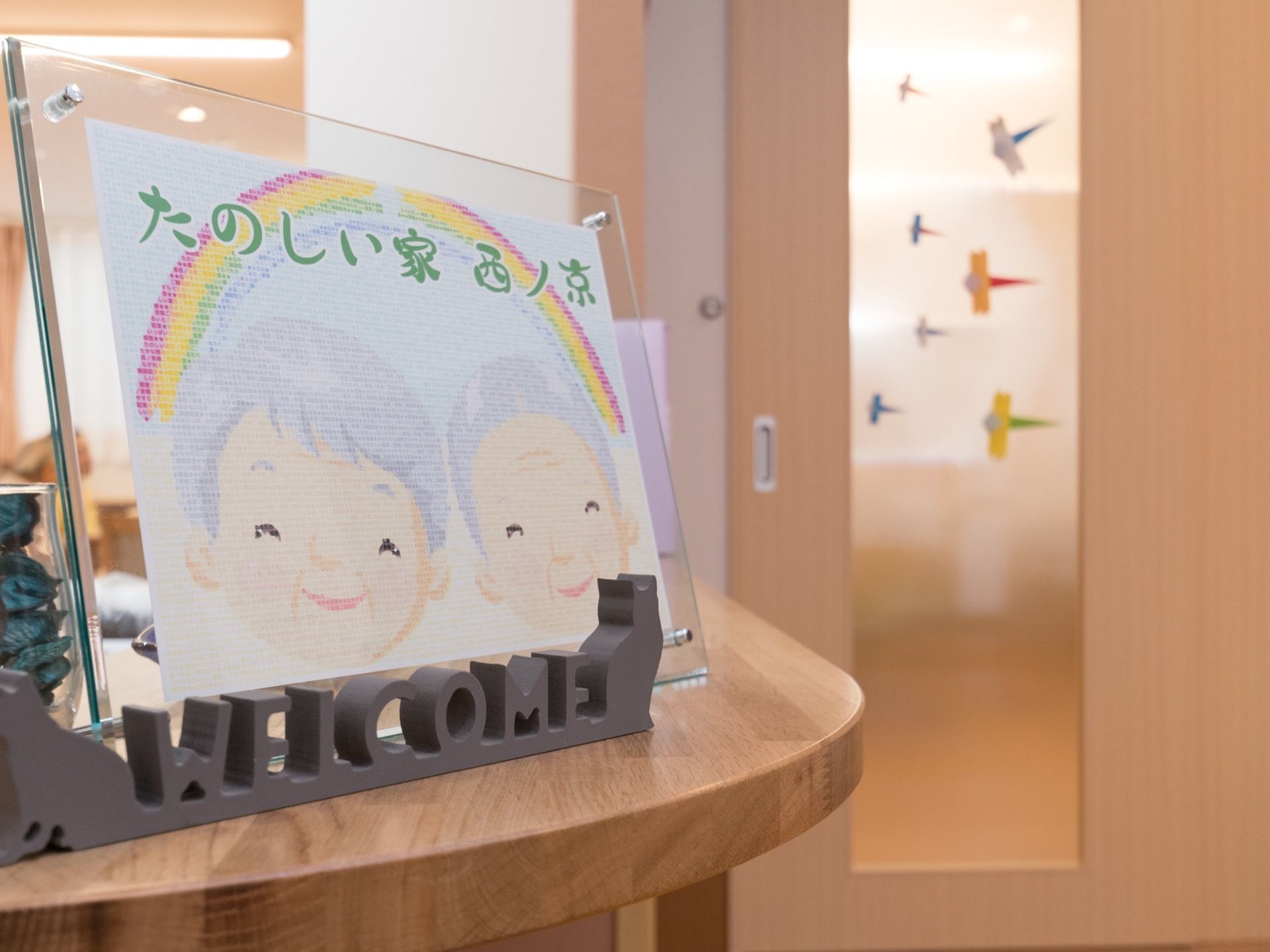 トンボの折り紙が飾られたドア、老人の絵が描かれた施設名の入ったイラストやwelcomeの置物が置かれているカウンター
