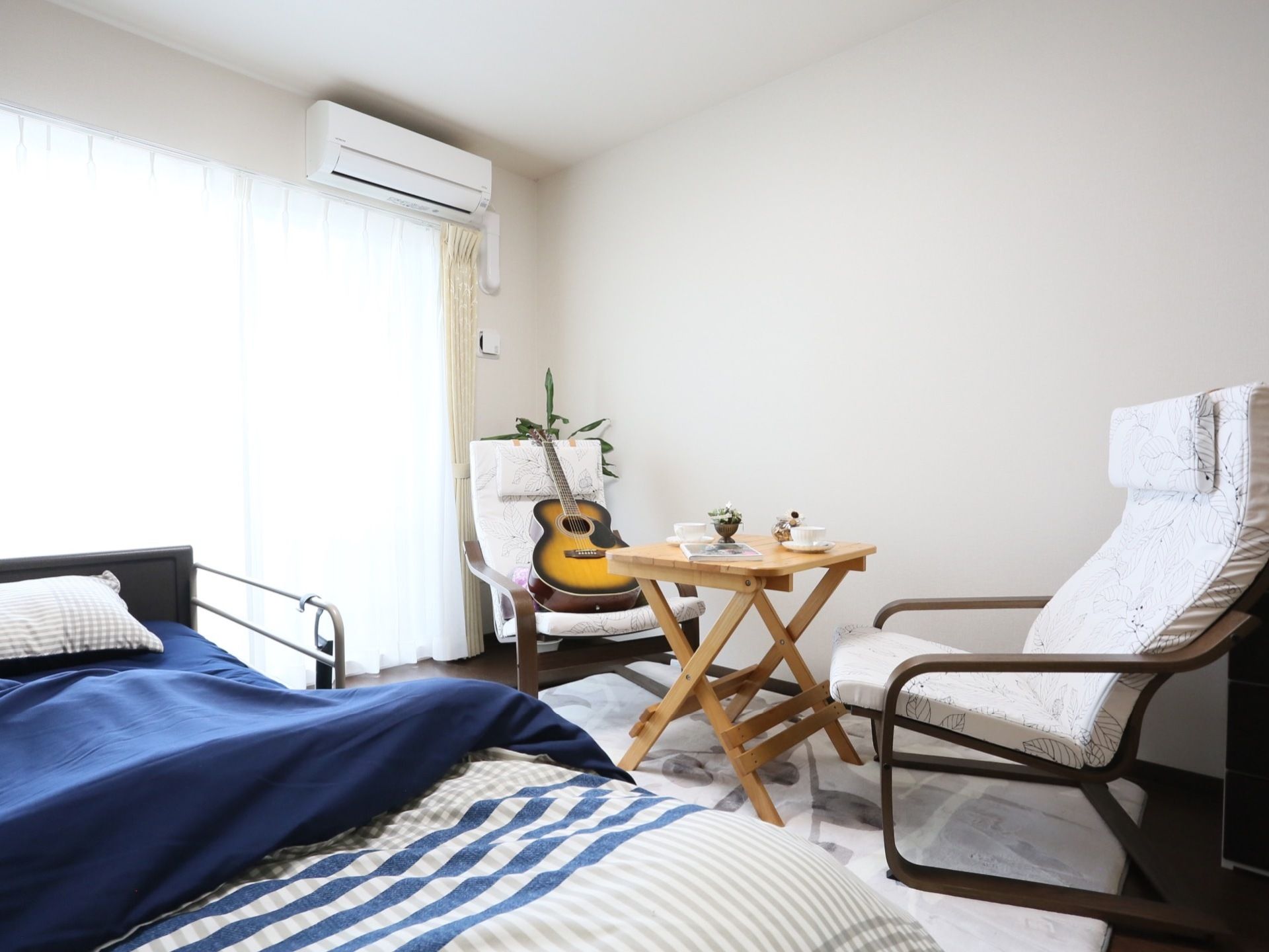 白を基調としたシンプルな居室。ベッドには手すりが取りつけられており、カーペットや観葉植物が設置されている。
