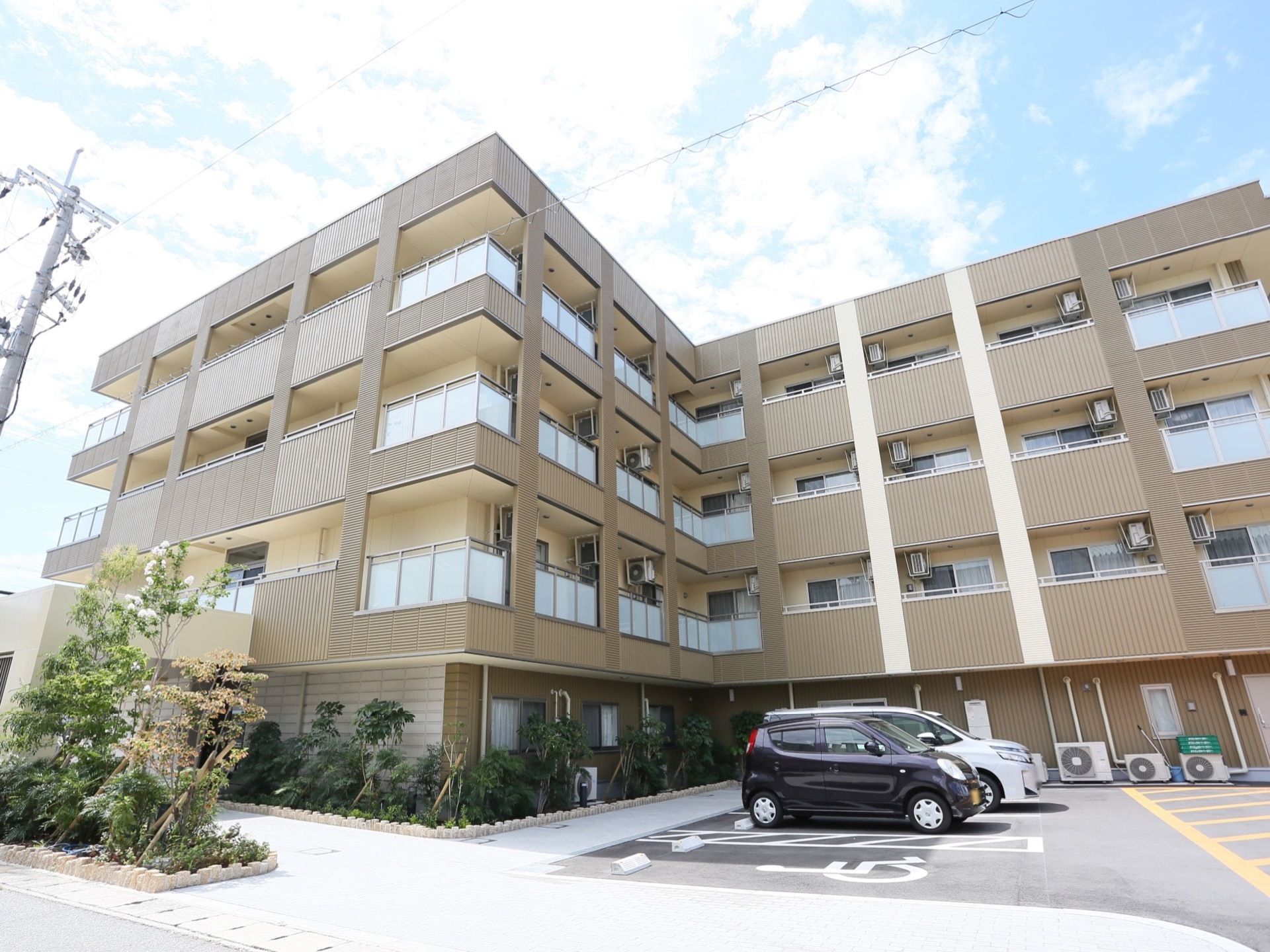 四階建てのマンションのような外観。周辺の花壇に植物が植えられており、駐車場は多数停められるようになっている。