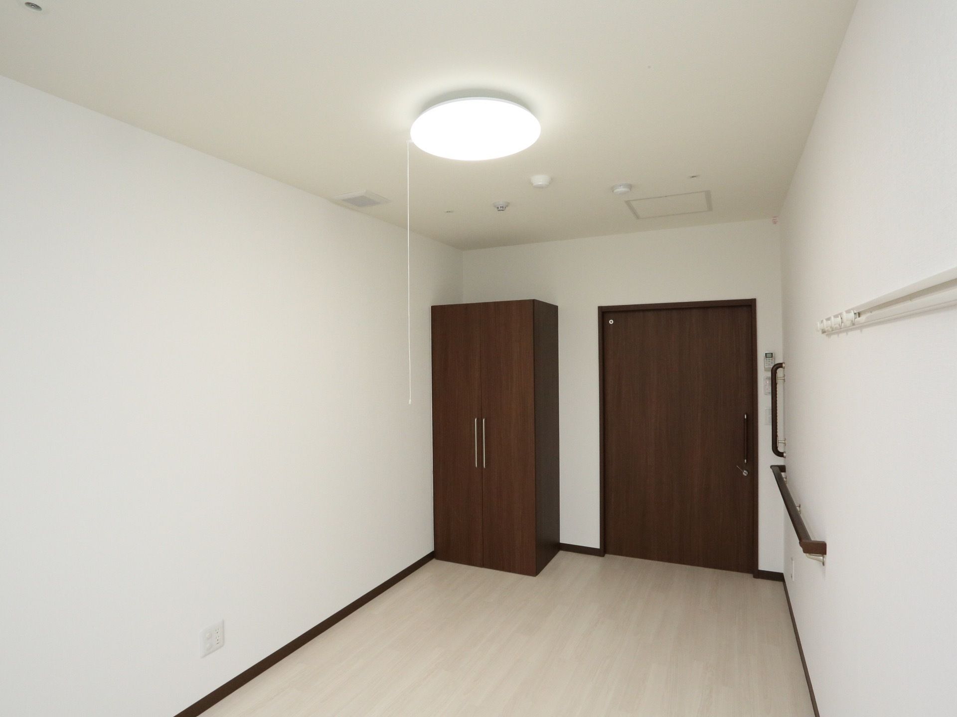 奥行きのある白い壁の部屋に、暗い木目調の引き戸の出入口がある。引き戸の左部には、観音開きの、木目調のクローゼットが置かれている。