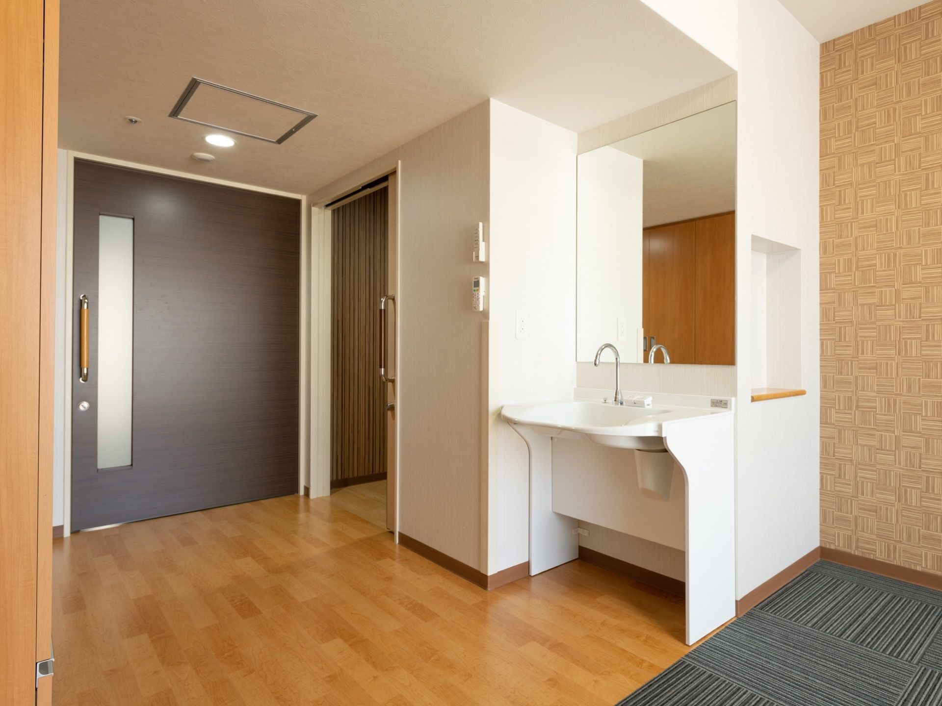 大きな鏡付きの白い洗面台がある。部屋の入口はスライドドアになっている。入口の斜向かいのスライドドアが開かれ、茶色いカーテンが見える。