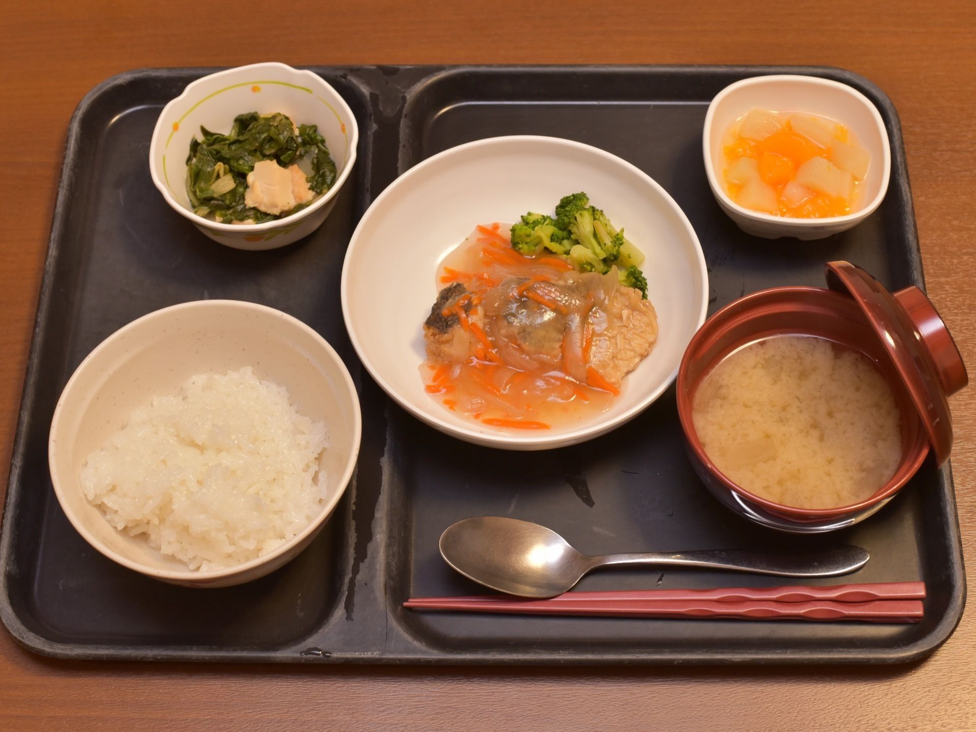 黒いお盆にセットされた食事。メインディッシュの魚のあんかけが中央にあり、味噌汁、小鉢とご飯のほか、デザートが添えられている。