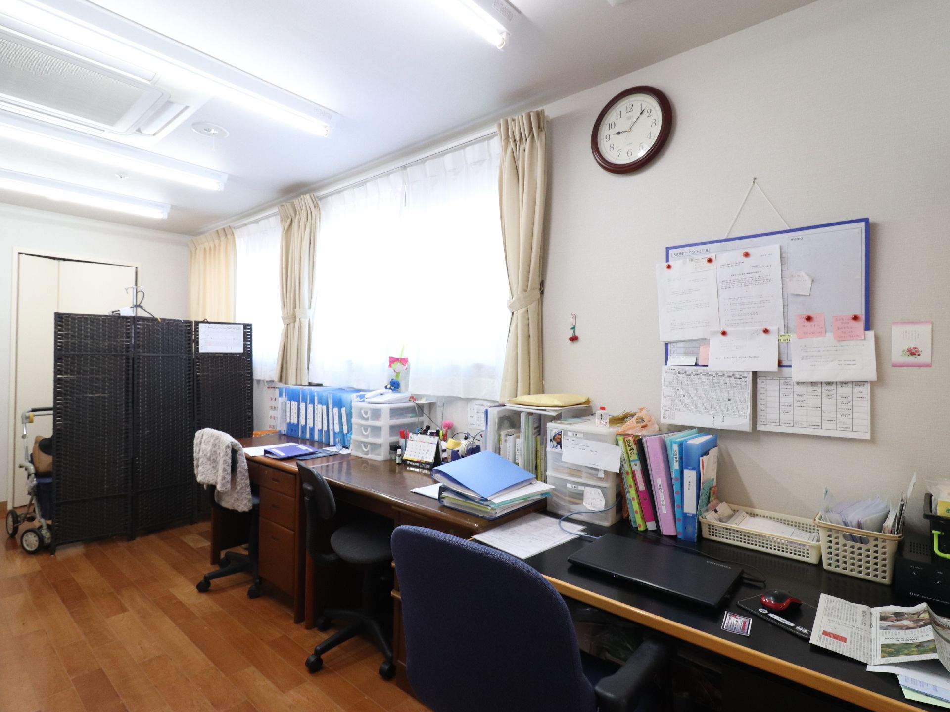 事務室は、作業机、椅子が置かれていて、机には色々な書類がある。壁には時計、ホワイトボードがあり、予定が分かるようになっている。