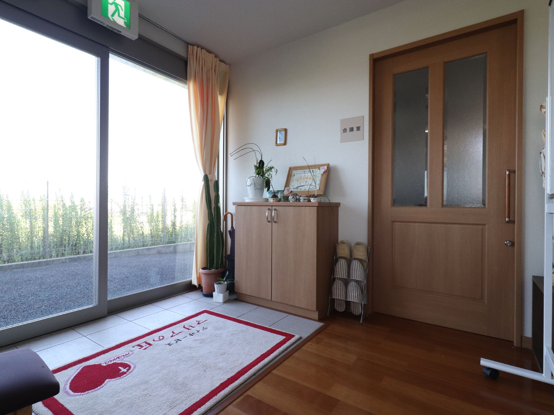 玄関ドアはガラス張りで、中が見えるようになっている。内側にはカーテンが束ねてある。床にはマットが敷かれ、靴箱やスリッパラックが置かれている。