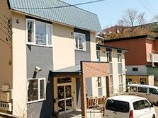 ベージュと黒に塗り分けられた、おしゃれな外観の２階建ての建物。玄関にはひさしがあり、ひさしの柱が門のようになっている。
