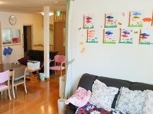 明るく広いリビングである。テーブルや椅子、ソファが置かれている。壁には入居者様がレクリエーションで描かれた絵が飾られている。