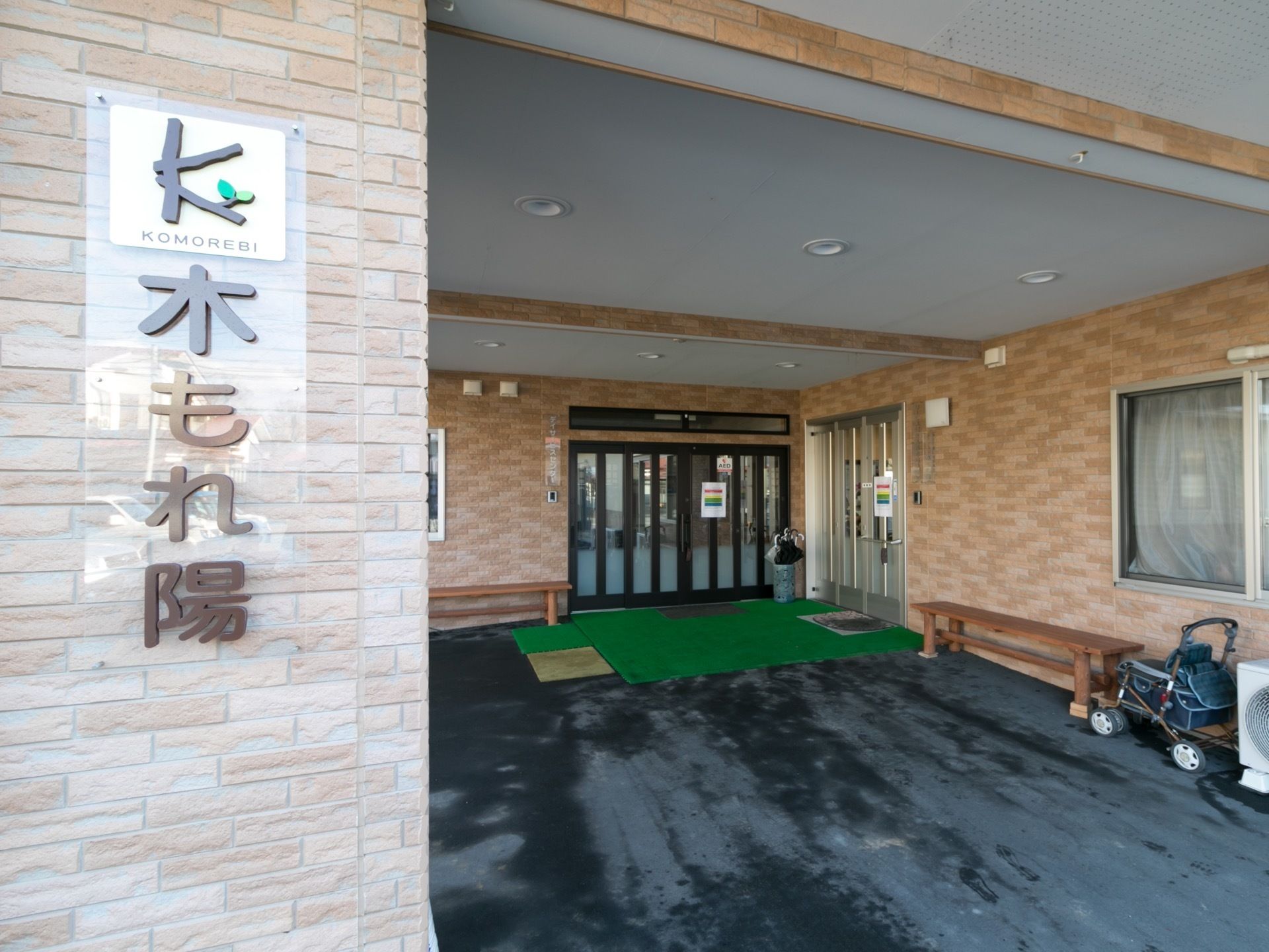 玄関の外観である。施設名の表記された柱の奥に玄関があり、屋根があるため悪天候でも安心である。出入口の前には緑のマットやベンチがある。