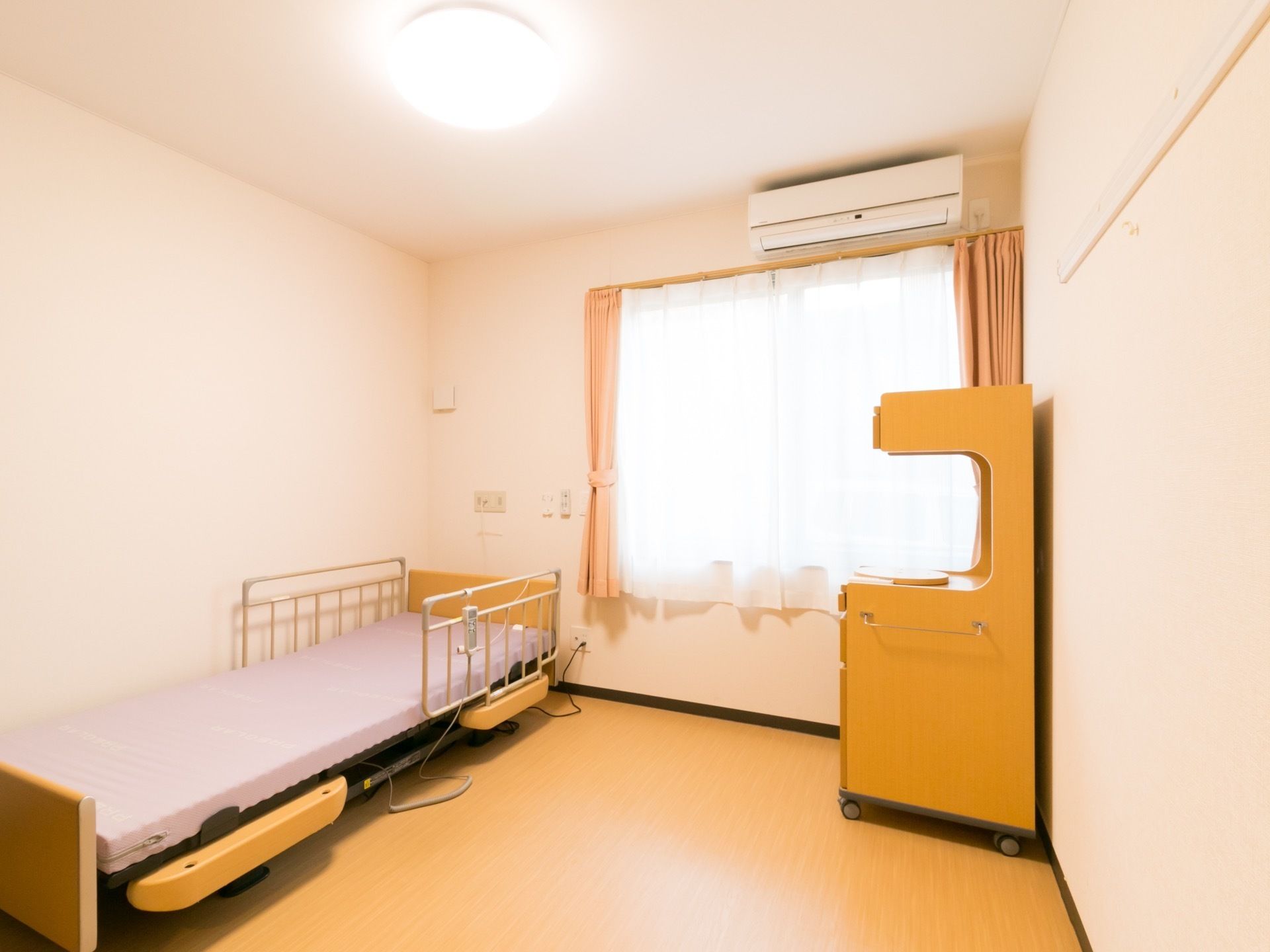 窓には2重のカーテンが掛けられエアコンが完備された居室である。マットレスが設置された介護用ベットとキャスター付き2段棚が設置されている。